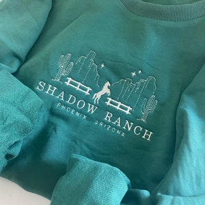 Könnte beinhalten: Ein tealgrüner Sweatshirt mit einem weißen gestickten Design eines Pferdes, das über einen Zaun mit Kakteen und Sternen springt. Der Text "SHADOW RANCH PHOENIX ARIZONA" ist unter dem Pferd gestickt.