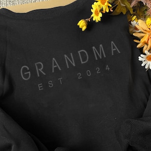 May include: Black sweatshirt with the text "GRANDMA EST 2024" printed on the front.