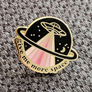 May include: A gold-coloured enamel pin with a black and pink design. The pin features a UFO flying over a planet with a crescent moon. The text around the planet reads "Give me more space".