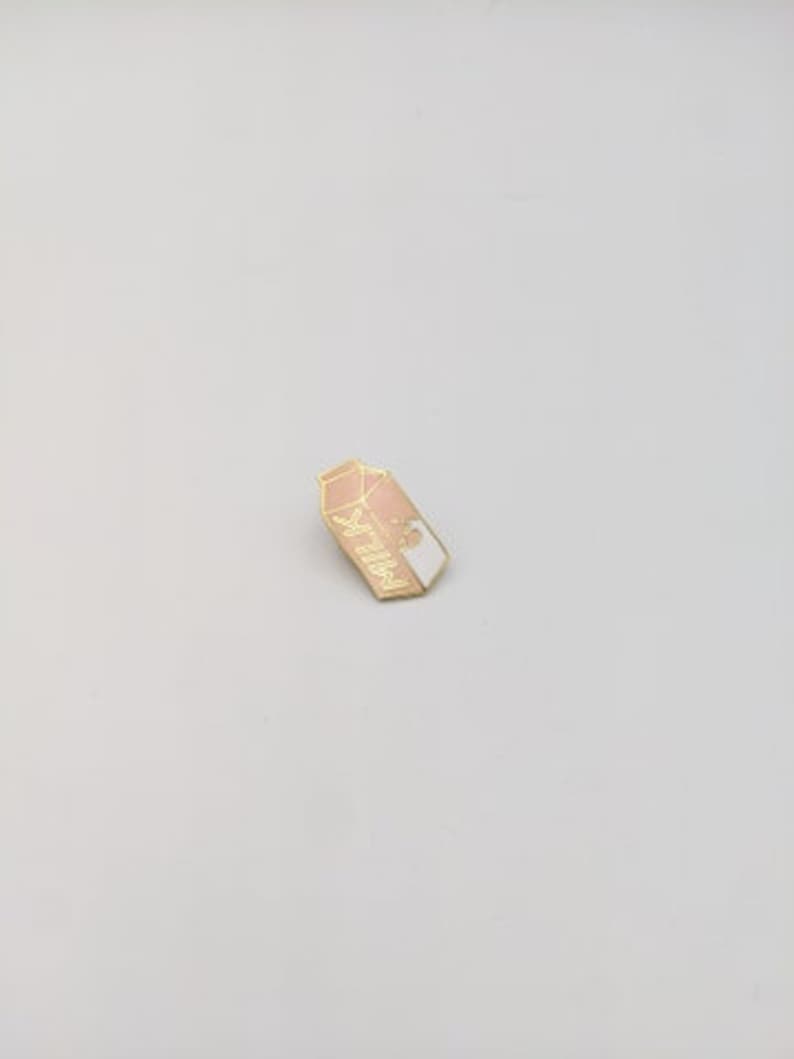May include: A gold-colored enamel pin shaped like a milk carton with the word "MILK" written on it. The carton is half pink and half white.