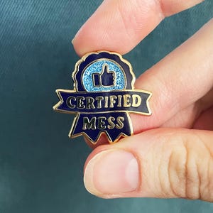 Pode incluir: Um alfinete dourado com um design azul e brilhante de polegar para cima. O alfinete diz "CERTIFIED MESS" em letras azul marinho. O alfinete é segurado entre os dedos contra um fundo verde-azulado.