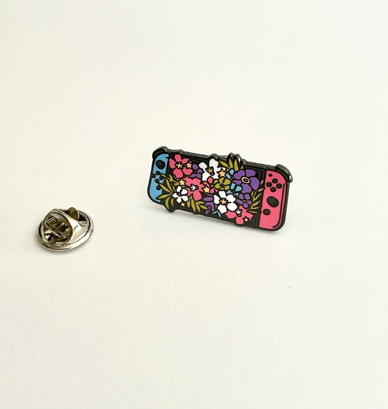May include: A black enamel pin featuring a Nintendo Switch console with a floral design. The console is pink and blue and the flowers are pink, purple, white, and green.