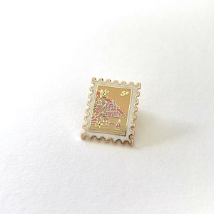 May include: Gold-colored enamel pin shaped like a postage stamp with a white background. The stamp features a small sailboat and a red and brown illustration of a building with palm trees. The stamp says "5c" in the upper right corner.