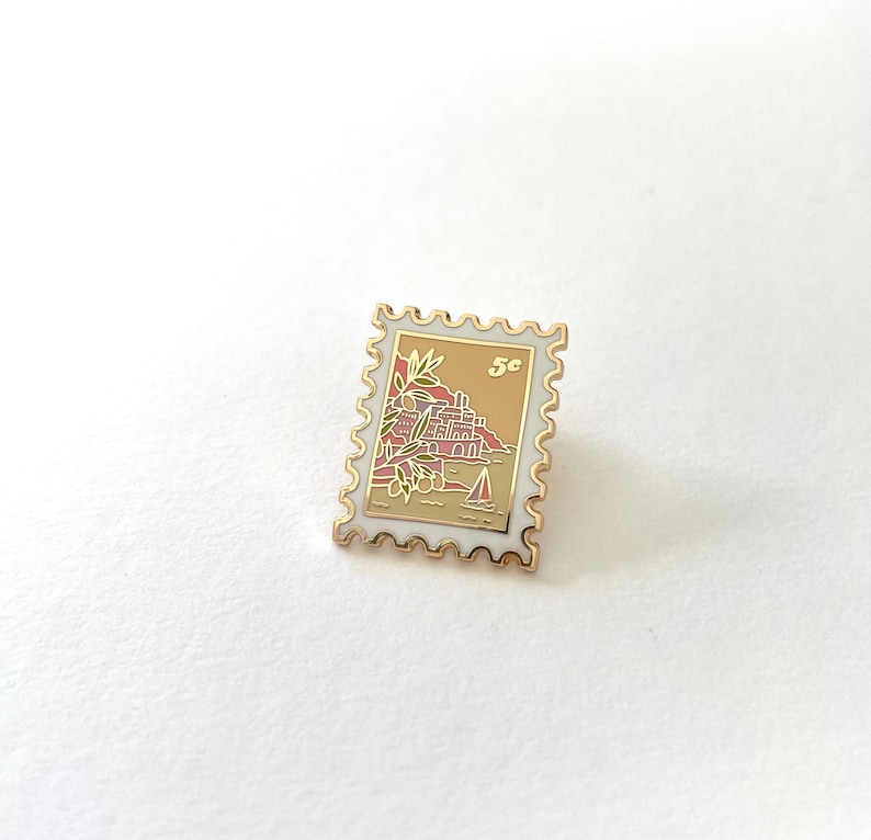 May include: Gold-colored enamel pin shaped like a postage stamp with a white background. The stamp features a small sailboat and a red and brown illustration of a building with palm trees. The stamp says "5c" in the upper right corner.