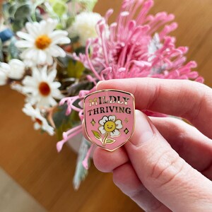 May include: A pink enamel pin with gold trim, featuring the words "Mildly Thriving" above a smiling daisy. The pin is held in a hand, with a blurred floral arrangement in the background.