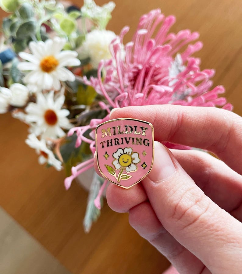 May include: A pink enamel pin with gold trim, featuring the words "Mildly Thriving" above a smiling daisy. The pin is held in a hand, with a blurred floral arrangement in the background.