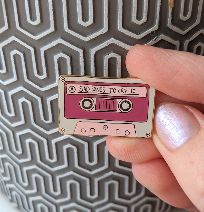 May include: A gold-colored enamel pin shaped like a cassette tape with the text "A SAD SONGS TO CRY TO" on the label. The cassette tape is pink and white.