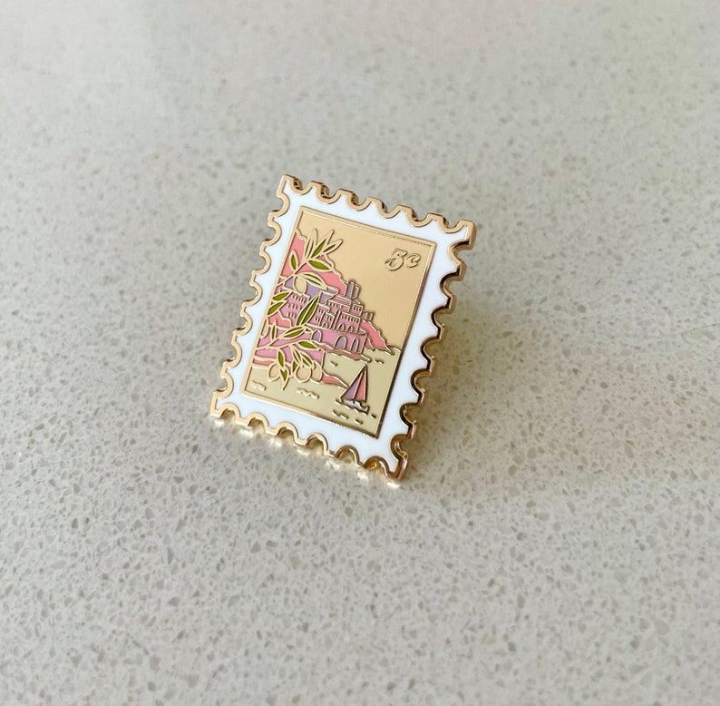 May include: Gold-tone enamel pin shaped like a postage stamp with a white border. The stamp features a pink and yellow illustration of a building on a cliff with a sailboat in the water. The stamp says "5c" in the upper right corner.
