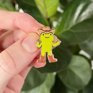 May include: A yellow enamel pin of a cartoon frog wearing a cowboy hat and boots. The frog has a gold outline and is holding its arms out. The cowboy hat and boots are a reddish-brown color.