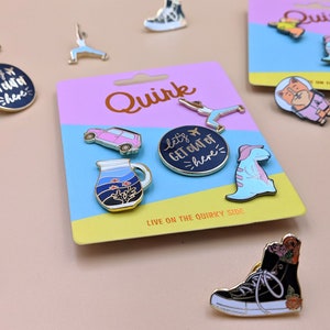 May include: A set of enamel pins featuring various designs, including a car, a jug, a dinosaur, a yoga pose, a cat, and a shoe. The pins are on a pink and blue card with the word "Quirk" printed on it. The text "Let's get out of here" is also on one of the pins.