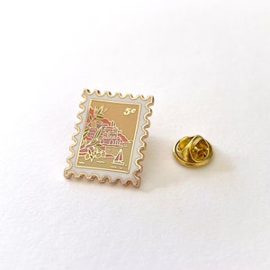 May include: Gold-toned enamel pin shaped like a postage stamp with a seaside scene and the text "5c".