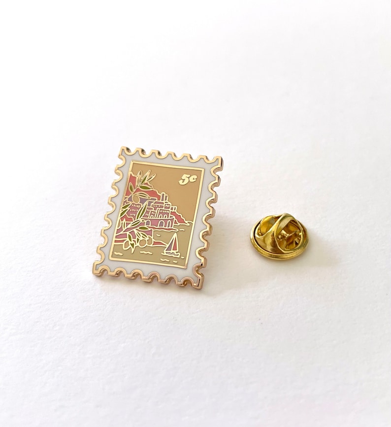 May include: Gold-toned enamel pin shaped like a postage stamp with a seaside scene and the text "5c".