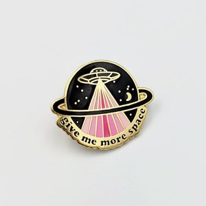 May include: A gold-colored pin with a black and pink design. The pin features a UFO with pink rays of light and the text "give me more space".