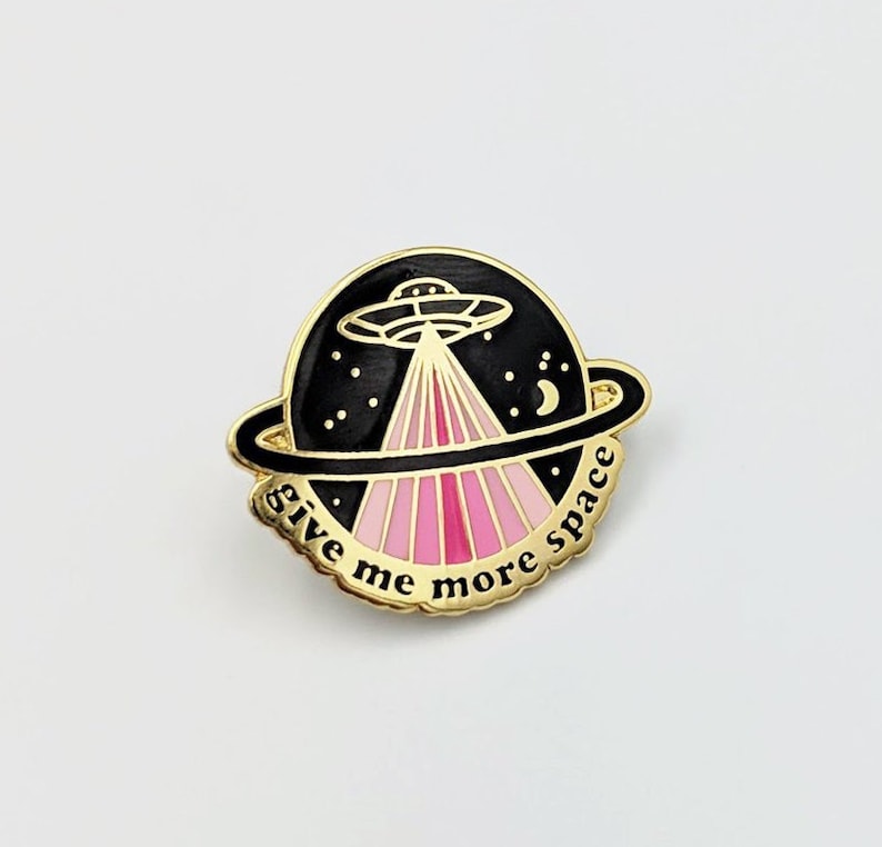 May include: A gold-colored pin with a black and pink design. The pin features a UFO with pink rays of light and the text "give me more space".