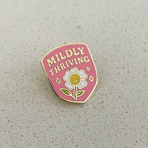 May include: A pink enamel pin with the words "Mildly Thriving" in gold lettering. The pin features a smiling daisy with a yellow center and white petals. The pin has a gold border and diamond accents.