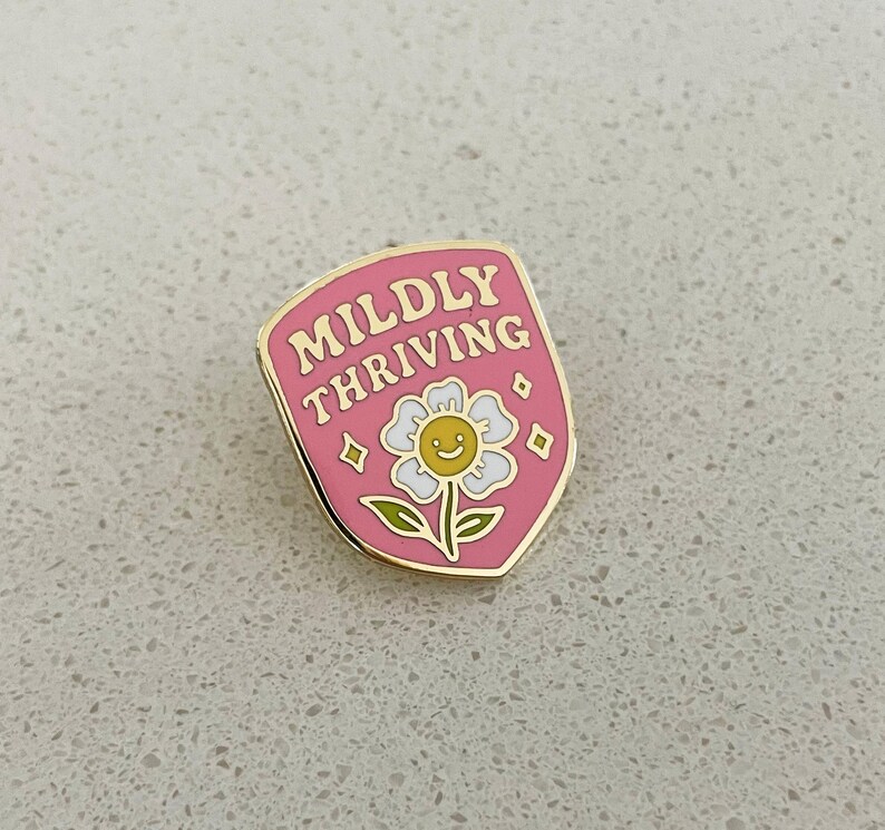 May include: A pink enamel pin with the words "Mildly Thriving" in gold lettering. The pin features a smiling daisy with a yellow center and white petals. The pin has a gold border and diamond accents.