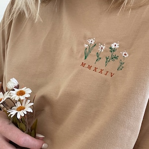 Könnte beinhalten: Ein hellbraunes T-Shirt mit einem gestickten Design aus vier weißen Blumen mit grünen Stielen und Blättern. Das Jahr "MMXXJV" ist unter den Blumen in braunem Garn gestickt.