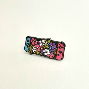 May include: A black and pink enamel pin featuring a Nintendo Switch console with a floral design. The flowers are pink, purple, white, and yellow.