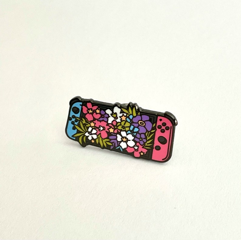 May include: A black and pink enamel pin featuring a Nintendo Switch console with a floral design. The flowers are pink, purple, white, and yellow.