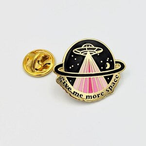 May include: Gold-tone enamel pin with a black and pink design of a UFO flying over a planet with the text "give me more space".