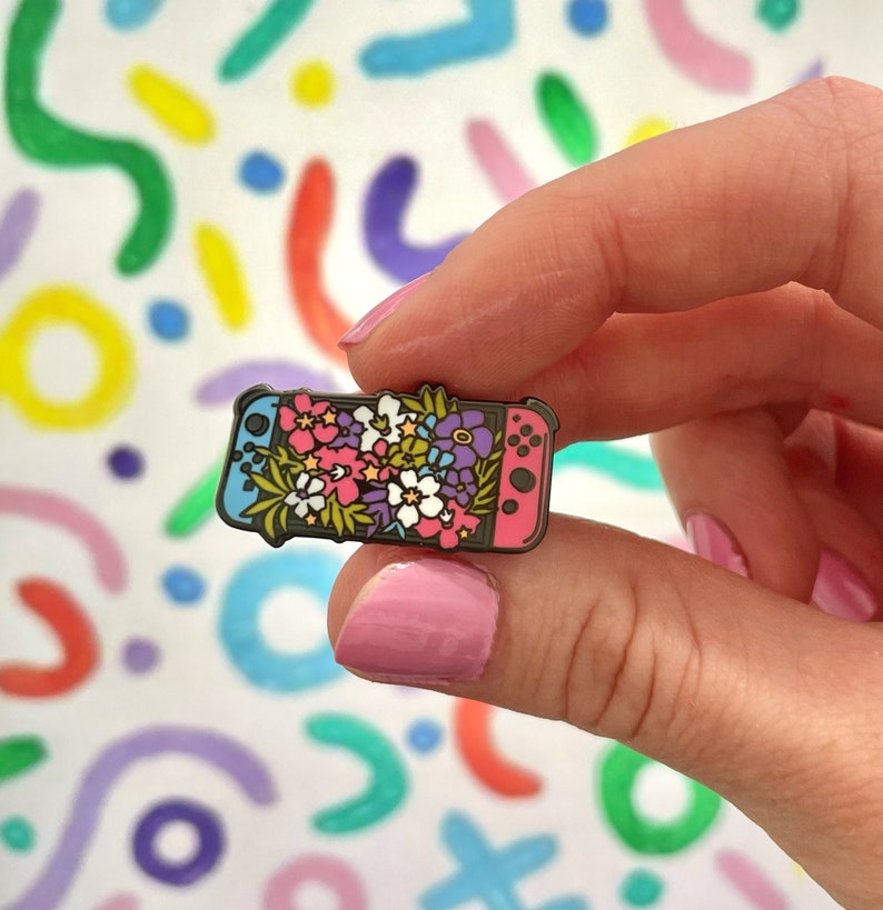 May include: A pink and blue enamel pin shaped like a Nintendo Switch console with a floral design. The flowers are pink, purple, white, and green.