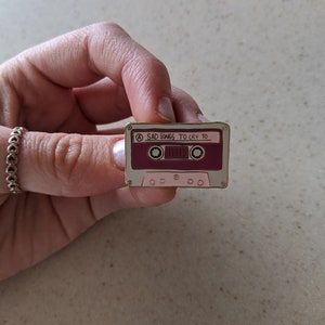 May include: A pink and white cassette tape enamel pin with the text "SAD SONGS TO CRY TO" on the front.