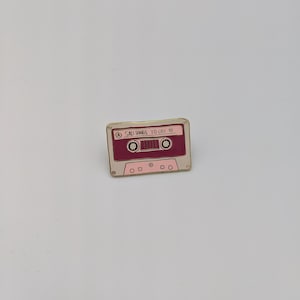 May include: Gold and pink enamel pin shaped like a cassette tape with the text "SAD SONGS TO CRY TO" on the front.