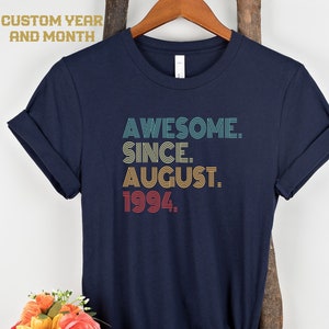 May include: A navy blue t-shirt with rolled sleeves. The shirt displays the text "AWESOME. SINCE. AUGUST. 1994." in teal, yellow, and red. The words "CUSTOM YEAR AND MONTH" are at the top. A bouquet of flowers is in the lower left corner.
