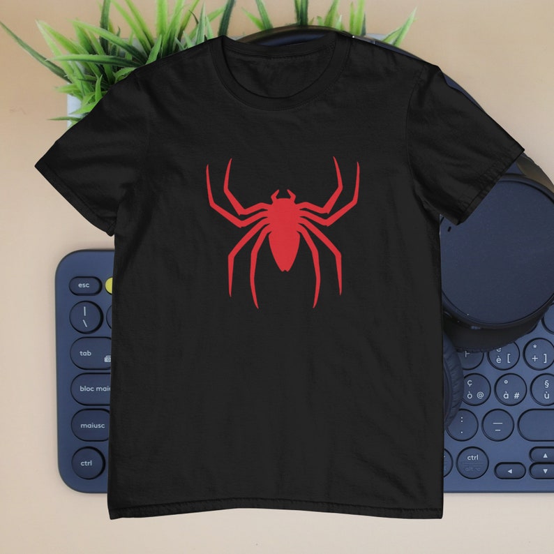Puede incluir: Una camiseta negra de manga corta con un gr&aacute;fico de ara&ntilde;a rojo s&oacute;lido y llamativo centrado en el pecho. El dise&ntilde;o de la ara&ntilde;a tiene ocho patas puntiagudas y distintas, adem&aacute;s de un cuerpo segmentado, creando un contraste n&iacute;tido con la tela.