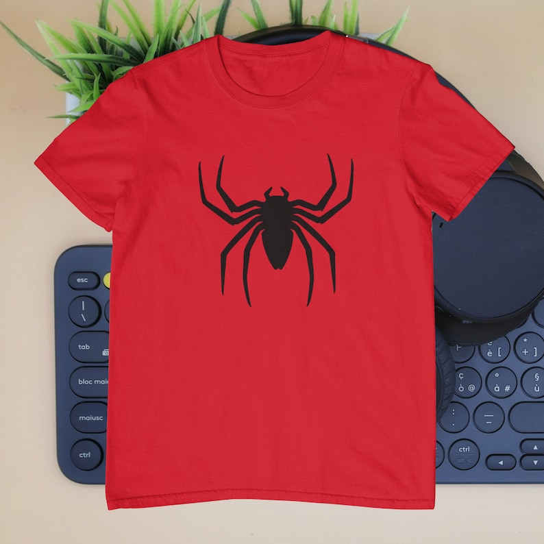 Puede incluir: Una camiseta roja de manga corta con un gr&aacute;fico negro grande y centrado de una ara&ntilde;a con ocho patas largas y puntiagudas y un cuerpo segmentado. La prenda tiene un cuello redondo cl&aacute;sico y est&aacute; confeccionada con un tejido liso y uniforme.