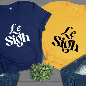 May include: Two t-shirts with the text "Le Sigh" printed on them. One shirt is navy blue with white text and the other is yellow with black text.