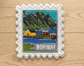 Peut inclure: Un écusson brodé rectangulaire en forme de timbre-poste représentant un paysage de montagnes, une étendue d'eau bleue et trois petites maisons jaunes et rouges. Le mot NORWAY est brodé en lettres capitales blanches en bas.