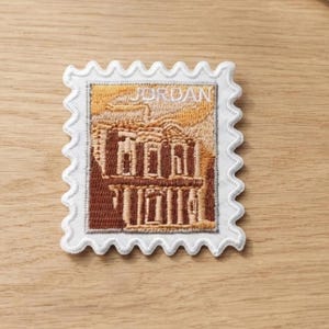 Puede incluir: Un parche bordado rectangular con forma de sello postal y borde festoneado blanco. El diseño muestra una escena arquitectónica en tonos marrones y beige con la palabra JORDAN bordada en hilo blanco en la parte superior del parche.
