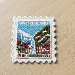 May include: A rectangular embroidered patch shaped like a postage stamp with a scalloped white border. The design features a Swiss mountain landscape with snow-capped peaks, a waterfall, and buildings, with the word SWITZERLAND in white text.