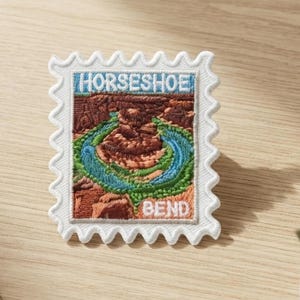 Può includere: Una toppa ricamata rettangolare a forma di francobollo raffigurante il paesaggio di Horseshoe Bend. Il design include formazioni rocciose marroni, un fiume blu serpeggiante e vegetazione verde. Il testo HORSESHOE BEND è ricamato in bianco.