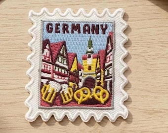 May include: A rectangular embroidered patch shaped like a postage stamp with a scalloped white border. The design features a German town scene with half-timbered buildings, a central tower, beer steins, and pretzels, with the text GERMANY above.