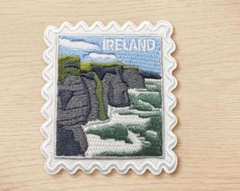 May include: A rectangular embroidered patch shaped like a postage stamp with a scalloped white border. The design features a landscape of dark gray cliffs, green grass, and white ocean waves under a blue sky, with the word IRELAND in white text.