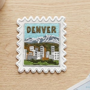 May include: A rectangular embroidered patch shaped like a postage stamp with scalloped edges. The design features the word DENVER in brown letters above a mountain range with snow-capped peaks, a city skyline, and a green foreground.
