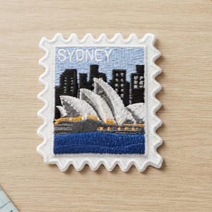 Può includere: Una toppa ricamata rettangolare a forma di francobollo che raffigura l'Opera di Sydney in bianco, uno skyline scuro e acqua blu. La parola SYDNEY è ricamata in bianco su uno sfondo di cielo azzurro con un bordo bianco smerlato.