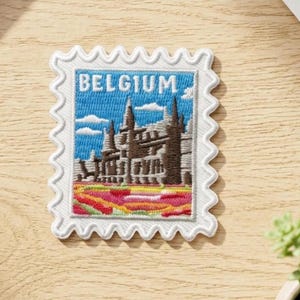 Può includere: Una toppa ricamata rettangolare con bordo bianco smerlato che raffigura un cielo blu con nuvole bianche, un edificio architettonico marrone e un campo di fiori rossi, gialli e rosa. La parola BELGIUM è ricamata in bianco nella parte superiore.