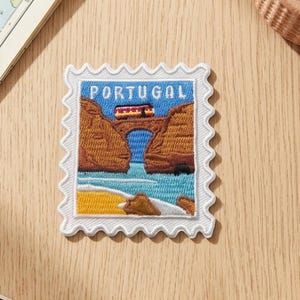 May include: A rectangular embroidered patch shaped like a postage stamp with a scalloped white border. The design features a landscape of brown cliffs, a blue ocean, a yellow sandy beach, and a small train crossing a bridge with the word PORTUGAL.