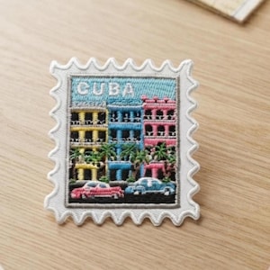 Puede incluir: Un parche bordado rectangular con forma de sello postal y borde blanco festoneado. El diseño muestra la palabra CUBA en letras blancas sobre edificios amarillos, azules y rosas, palmeras verdes y dos coches antiguos.