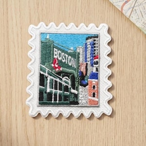 May include: A square embroidered patch with a scalloped white border featuring a detailed depiction of a green stadium wall with the word BOSTON in white letters, a red logo, and surrounding city buildings under a bright blue sky.