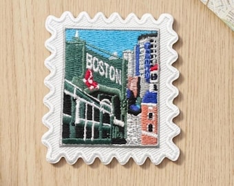 Puede incluir: Un parche bordado cuadrado con un borde blanco festoneado que presenta una representación detallada de un muro de estadio verde con la palabra BOSTON en letras blancas, un logotipo rojo y edificios urbanos bajo un cielo azul.