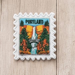 May include: A rectangular embroidered patch shaped like a postage stamp with a white scalloped border. The design features a waterfall cascading between orange cliffs and green pine trees, with the word PORTLAND printed in dark blue at the top.