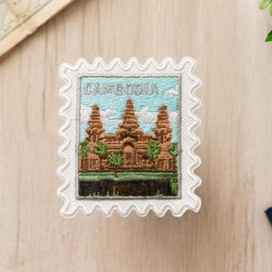 Può includere: Una toppa ricamata quadrata a forma di francobollo con bordo bianco smerlato. Il disegno raffigura il tempio di Angkor Wat in Cambogia in tonalità marroni e verdi su un cielo azzurro con la parola CAMBODIA scritta nella parte superiore.