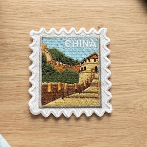 May include: A rectangular embroidered patch shaped like a postage stamp featuring a landscape of the Great Wall of China. The design includes green hills, a tan stone wall, and a light blue sky with the word CHINA embroidered in white thread.