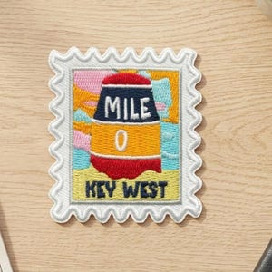May include: A rectangular embroidered patch shaped like a postage stamp with a scalloped white border. The design features a colorful illustration of the Key West Mile 0 marker buoy in red, yellow, and blue, with the text MILE 0 and KEY WEST.