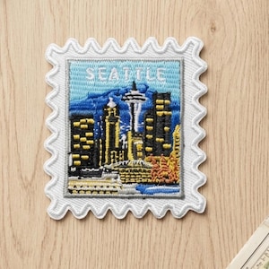 May include: A rectangular embroidered patch shaped like a postage stamp with a scalloped white border. The design features the Seattle skyline with the Space Needle, dark buildings with yellow windows, and blue mountains under the text SEATTLE.