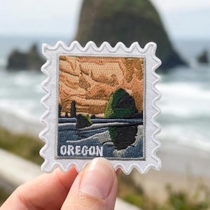 May include: A rectangular embroidered patch shaped like a postage stamp with a scalloped white border. The design features a landscape with a large dark green rock formation, blue ocean waves, and a textured orange sky. The word OREGON is printed.